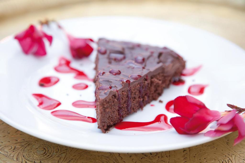 Šokolādes kūka, pasniegta uz balta šķīvja ar dekoratīviem ziediem un sarkanām mērces pēdām.