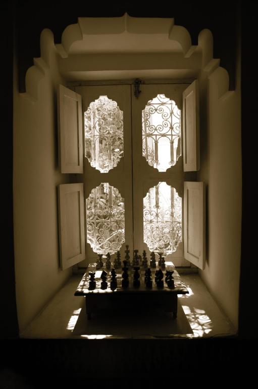 Šahs uz galda, kas novietots logā ar atvērtiem durvīm, caur kurām ieplūst gaisma. Logi rotāti ar izsmalcinātiem metāla ornamentiem, radot elegantu atmosfēru.