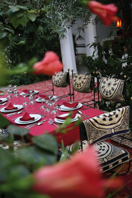 Eleganti uzklāta galda kompozīcija ar sarkanu galda segumu un dekoratīvām krēslu spilvenu apdarēm, iestādīta zaļajā dārzā. Galda klājumā redzami trauki, glāzes un salvetes, radot svinīgu atmosfēru.