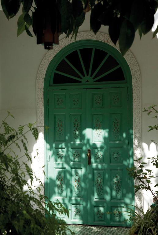 Zaļas durvis ar greznu apdari, kas ieskautas ar zālēm un ēnu spēlēm, radot mierīgu un aicinošu atmosfēru.