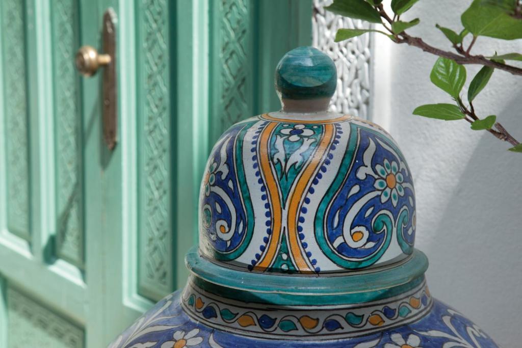 Tradicionāla keramikas trauka augšdaļa ar krāsainiem ornamentiem, fona zils un zaļš, ar dekoratīvu durvju daļu aizmugurē.