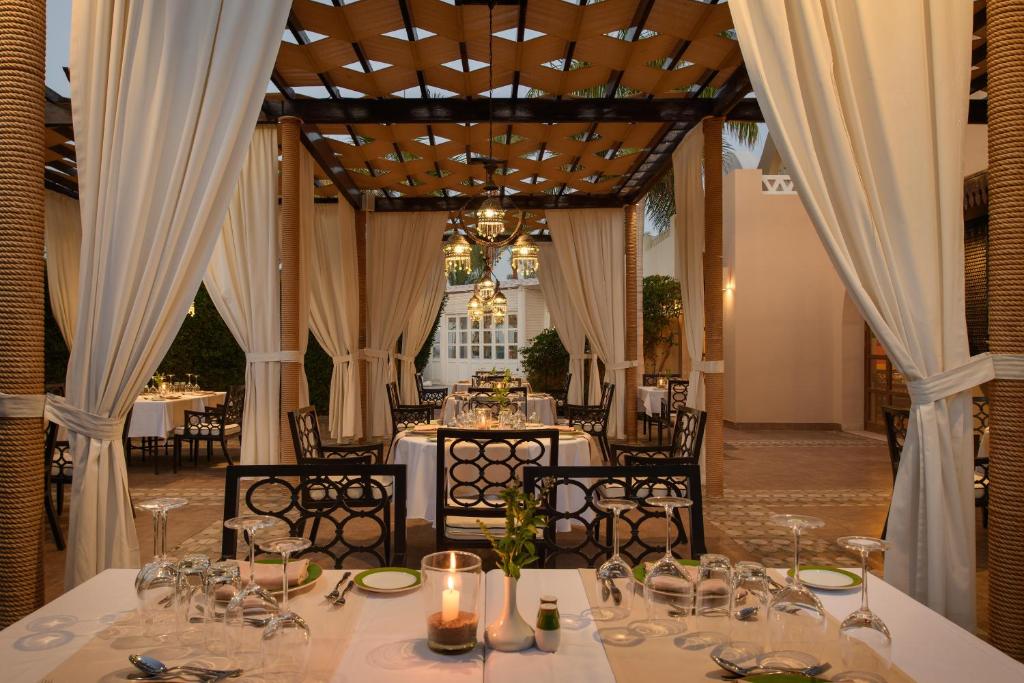 Elegants restorāns ar āra terasi, kurā ir balti galdauti un skaisti galda piederumi, apgaismots ar stilīgām laternām. Dāvā relaksējošu atmosfēru ar zaļumiem fonā.