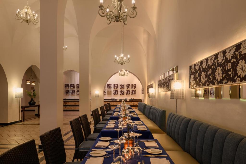 Elegants restorāna interjers ar garu galdu, dekorētu ar zilu galda pārklāju un elegantiem galda piederumiem. Gaismas ķermeņi un mākslinieciskas sienas piešķir atmosfēru.