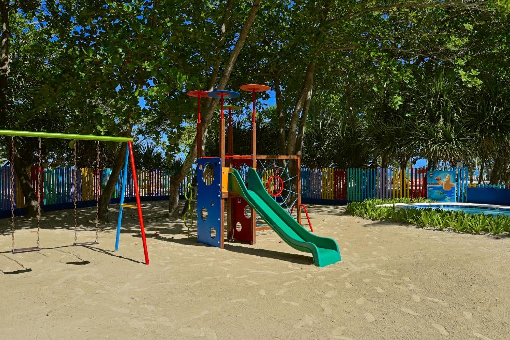 Krāsains bērnu laukums ar šūpolēm un slidkalniņu, apjozts ar koka žogu un zaļiem augiem. Ideāla vieta bērnu spēlēm un atpūtai.