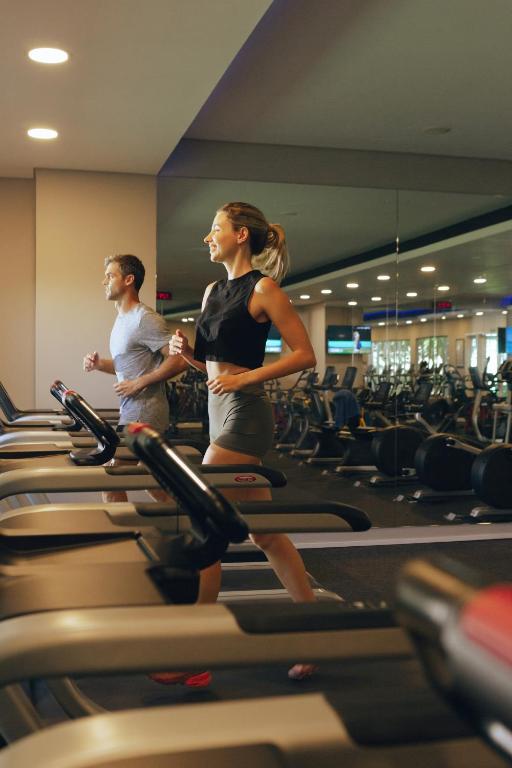 Divi cilvēki skrien uz skrejceliņiem modernā sporta zālē, ar spoguļiem un fitnesa aprīkojumu fonā. Gaisma ir silta un patīkama, radot motivējošu atmosfēru.