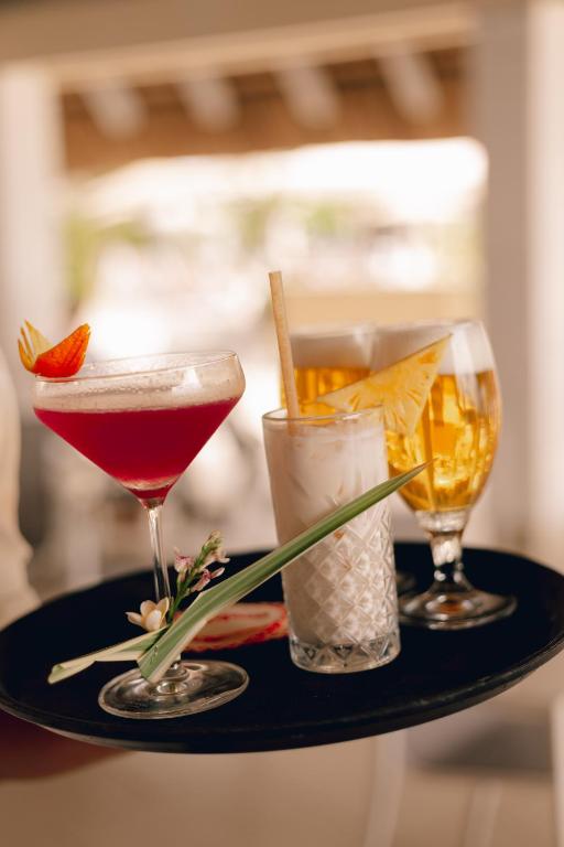 Trīs kokteiļi uz melna paplātes, tostarp sarkans kokteilis ar augļu rotājumu, balts kokteilis ar ananāsu un dzidrs alus.