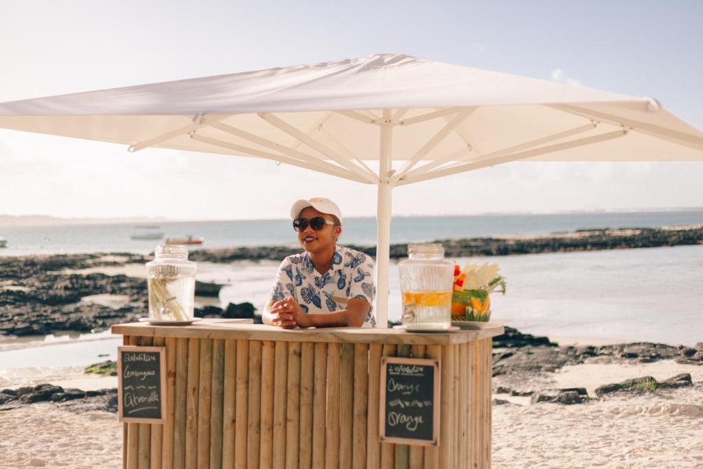 Pludmales stendā, zem saulessarga, stāv cilvēks, kas piedāvā dzērienus, ieskaitot apelsīnus un citronus. Fons ir pludmale ar jūru un akmeņiem.