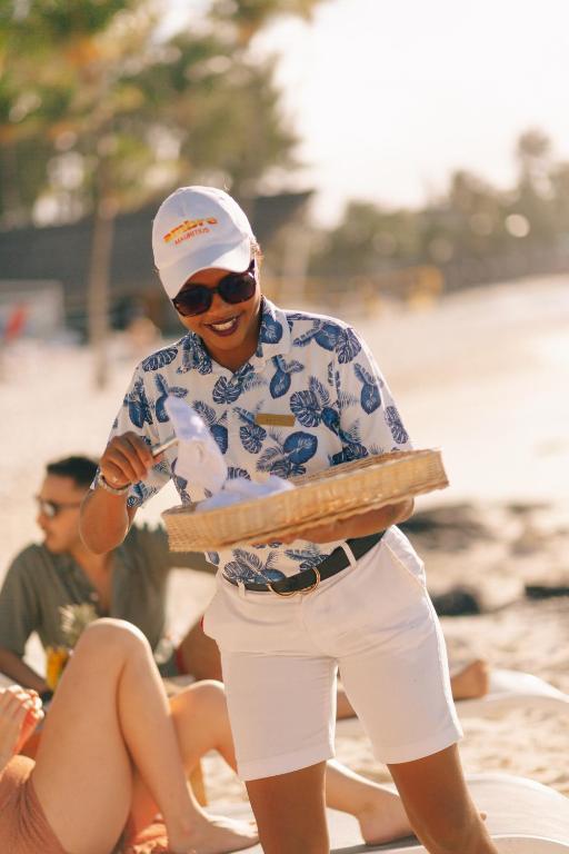 Sieviete pludmalē, tērpta ziedu raksta krekliņā un baltās šortos, pasniedz ēdienu no groza. Aiz viņas redzami atpūtušies cilvēki, baudot sauli un pludmales atmosfēru.