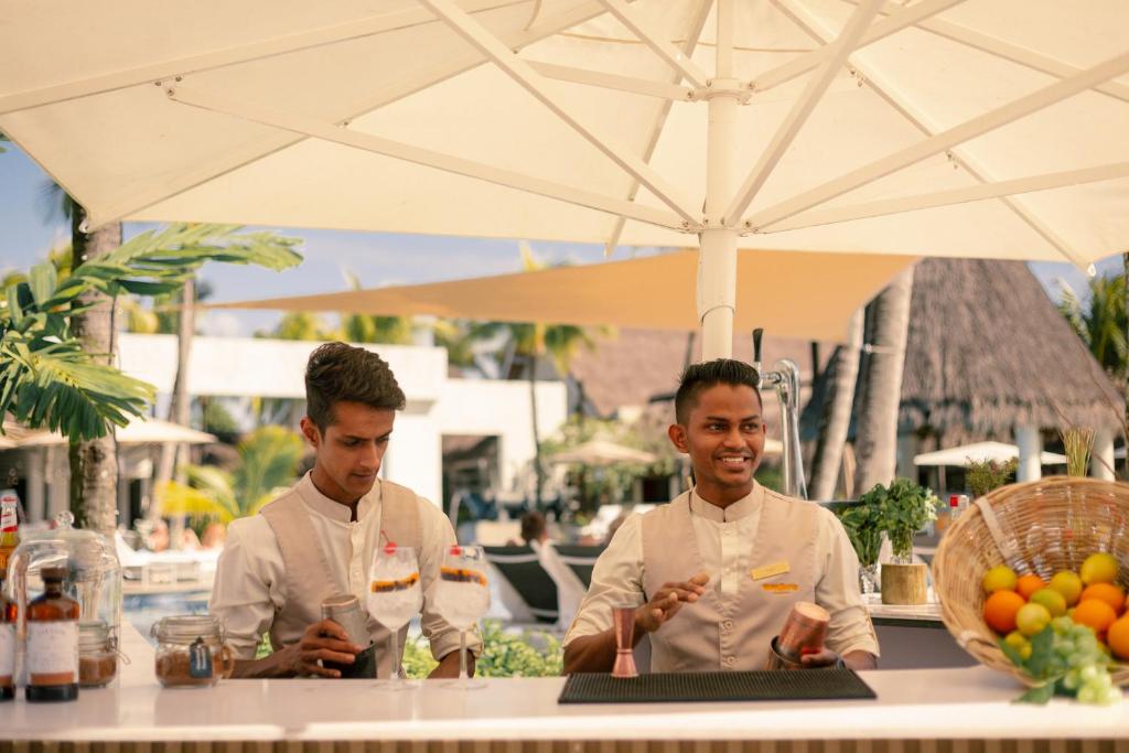 Jauna bāra atmosfēra, kur divi viesmīļi pasniedz dzērienus, apkalpojot viesus tropiskā vidē. Uz galda ir pieejami augļi un dzērienu sastāvdaļas.