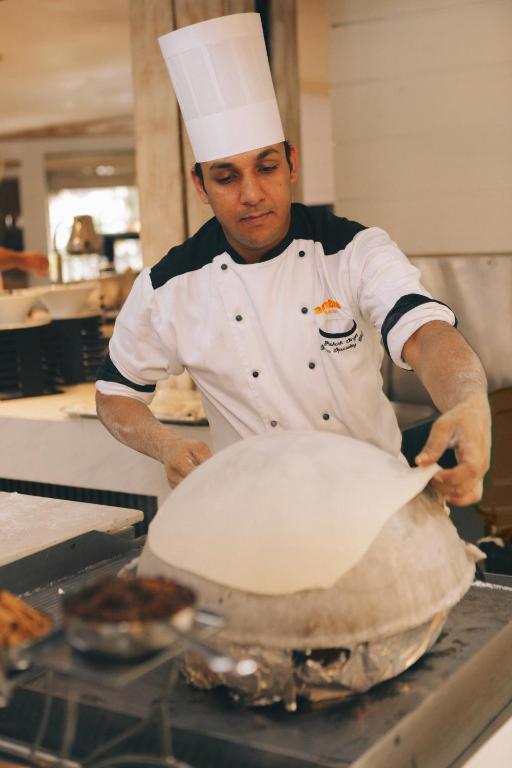 Šefpavārs, ģērbies balta uniformā, uzmanīgi noliek mīklas kārtu uz lielas katla vāka, gatavojot ēdienu restorānā. Fons ir mājīgs un profesionāls, radot kulinārijas atmosfēru.