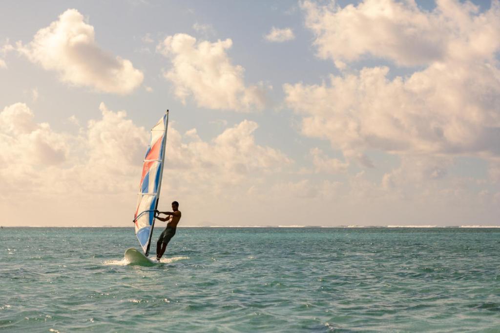 Vindsērfotājs, kas brauc pa viļņiem saulainā dienā, ar krāsainu buru un debesīm, kas klātas ar mākonīšiem.