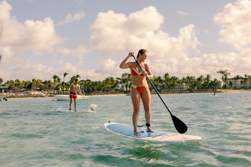 Divi cilvēki, kas nodarbojas ar stāvot peldēšanu, baudot saulainu dienu pie pludmales. Ūdens ir skaidrs, un fons ir zaļi palmu koki un viesnīcas.