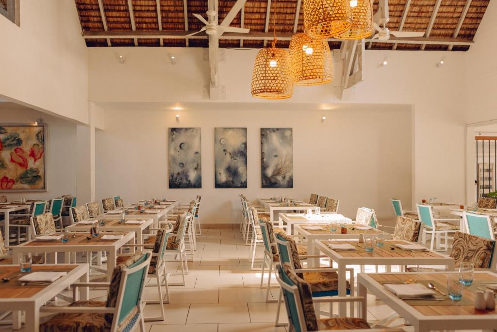 Moderns restorāns ar gaišu interjeru, koka griestiem un elegantiem galdiem, kas izkārtoti ar krāsainām krēsliem. Uz sienām redzami mākslas darbi, kas piešķir telpai radošu atmosfēru.