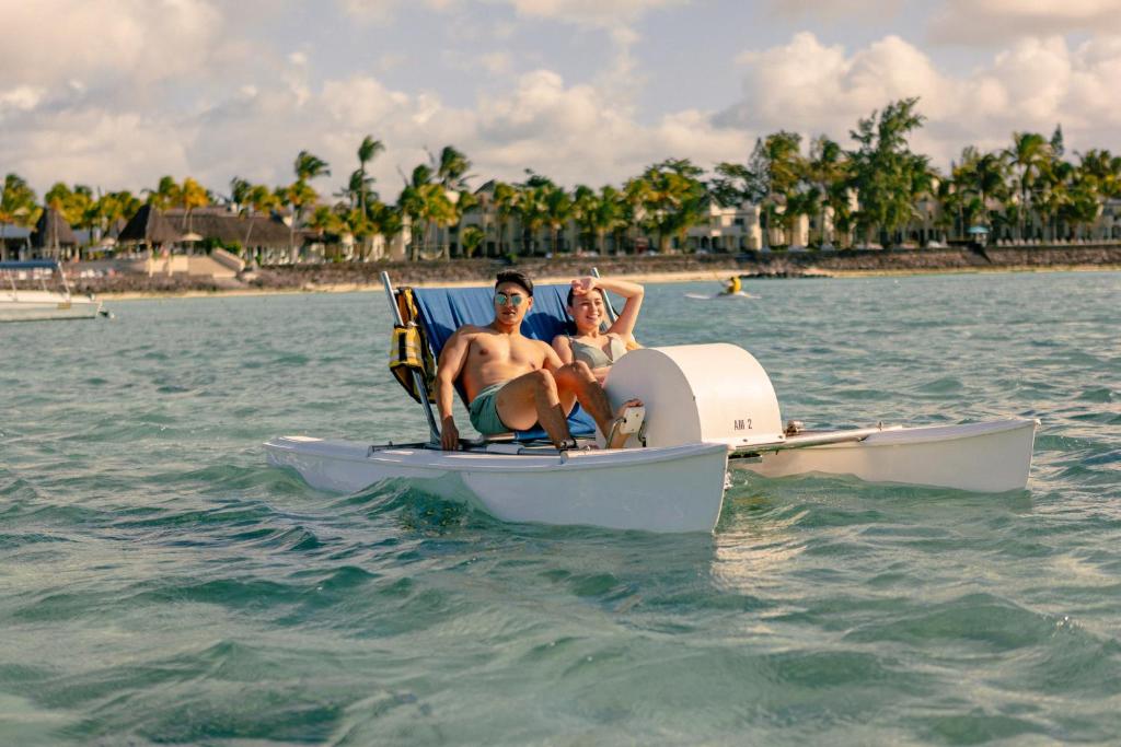 Pāris relaksējas uz katamarāna, izbaudot saulainu dienu pie ūdens ar palmām fona. Ūdens ir dzidrs, un apkārt redzami citi laivas.