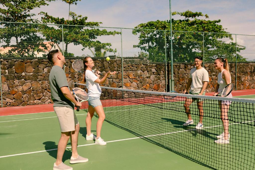 Jaunieši spēlē tenisu uz laukuma, izskatās priecīgi un aktīvi. Viens no spēlētājiem sagatavo bumbu, kamēr pārējie viņu vēro un smaidot sarunājas.
