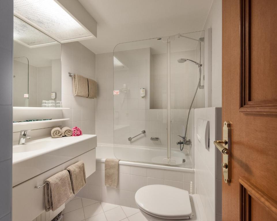 Mūsdienīgs un ērts vannas istabas interjers ar baltām flīzēm, vannu un dušas kabīni, kā arī elegantiem piederumiem. Vannas istabā redzami dvieļi un dekoratīvs zieds.