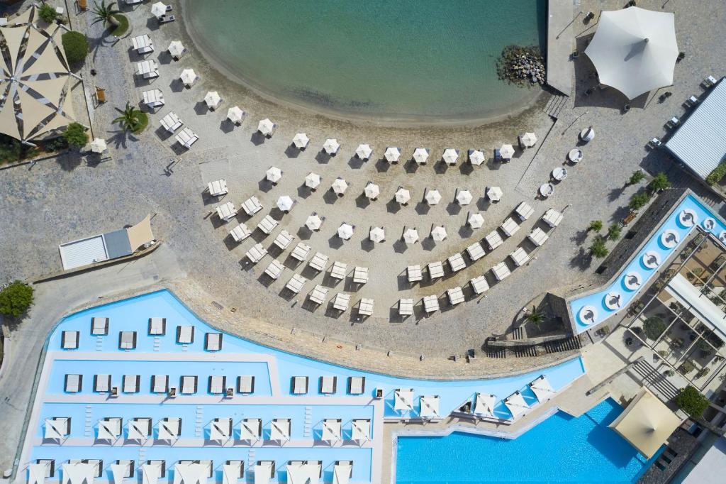 Viesnīcas komplekss ar baseinu un pludmali, kurā redzami sauļošanās krēsli un saulessargi. Skats no augšas, atklājot relaksējošu atmosfēru un tuvumu ūdenim.