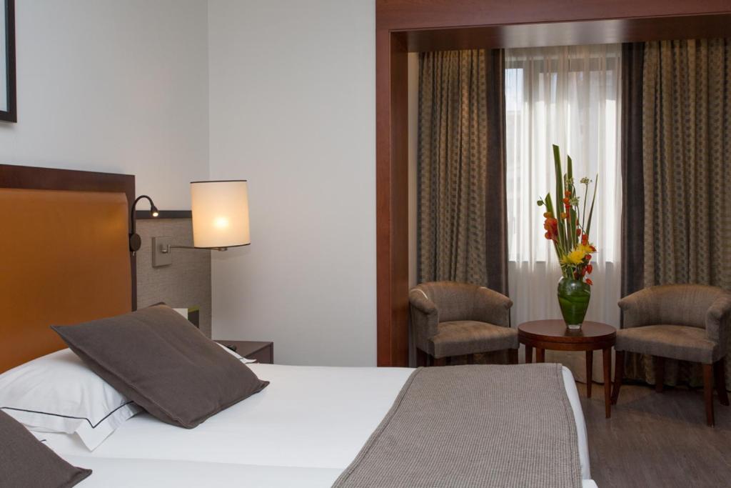 Mūsdienīgs viesnīcas numurs ar ērtu gultu, dīvāniem un dekoratīviem ziediem. Gaisma plūst caur logu, radot patīkamu atmosfēru.