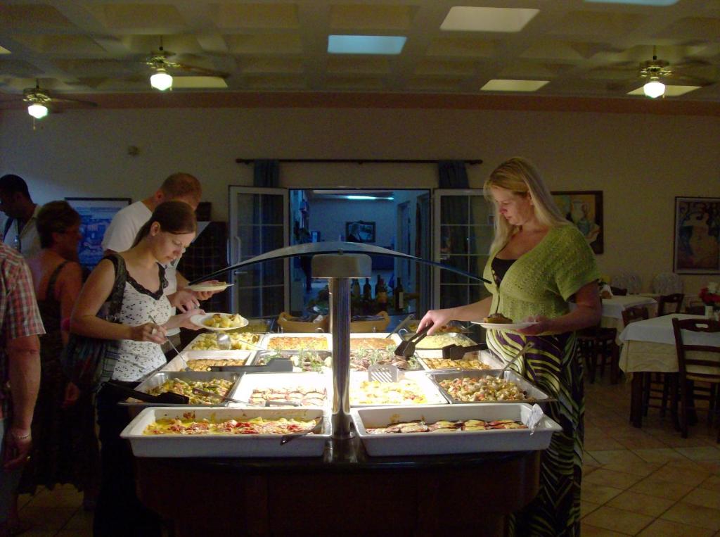 Buffet ar dažādām ēdienu izvēlēm, apmeklētāji ņem ēdienu, kamēr citi sēž pie galdiem.