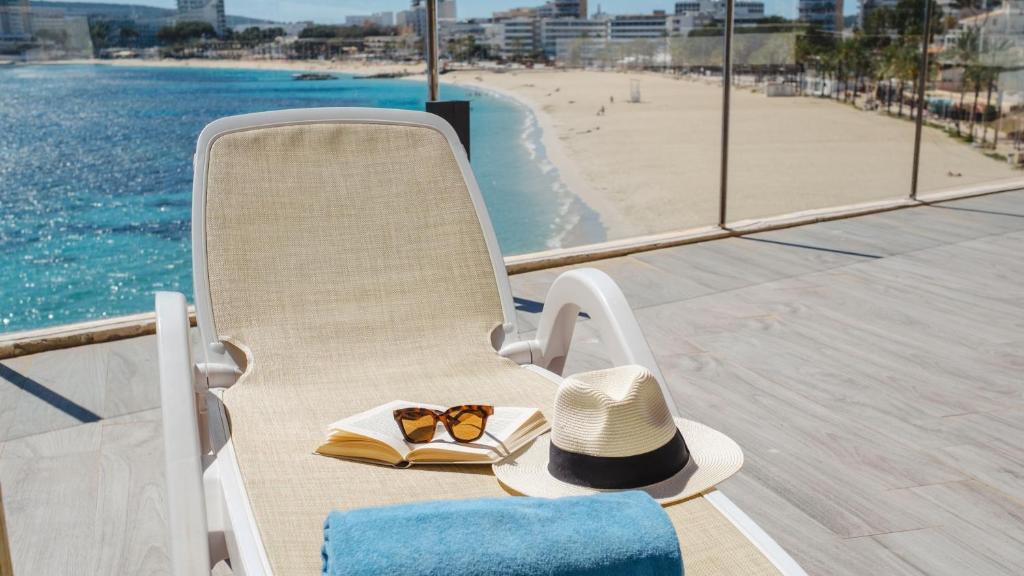 Saulaina atpūtas zona ar sauļošanās krēslu, grāmatu, saulesbrillēm un cepuri, fonā skats uz pludmali un jūru. Ideāla vieta relaksācijai un brīvā laika pavadīšanai.