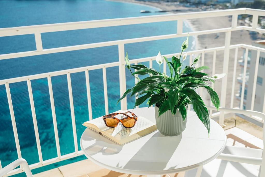 Saulaina terase ar galdu un krēslu, uz kura atrodas grāmata, saulesbrilles un zieds. Fons ir jūras skats ar smiltīm.