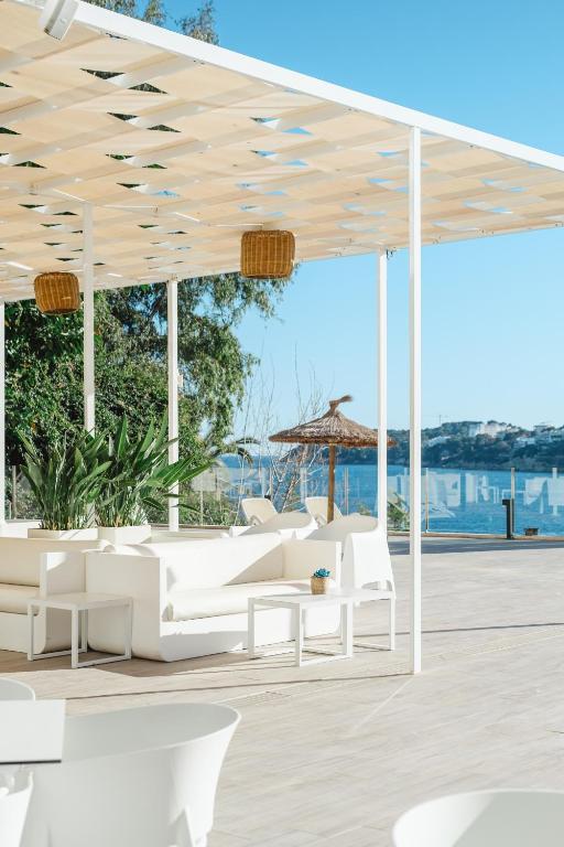 Mūsdienīgs terases dizains ar baltām mēbelēm un zaļu veģetāciju, kas atrodas pie ūdens. Ideāla vieta relaksācijai saulainā dienā.