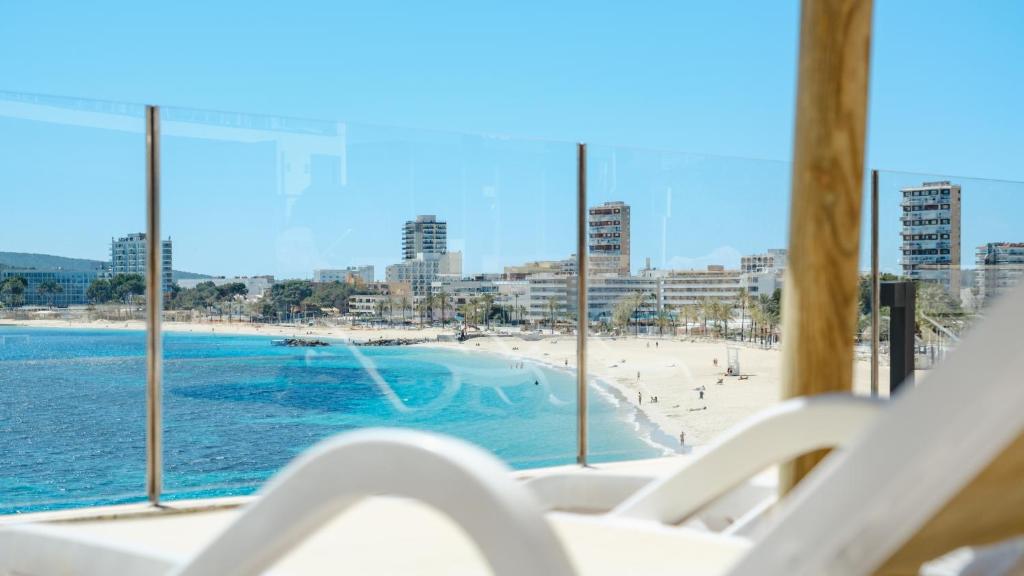 Skats uz pludmali ar skaidru zilo ūdeni un pilsētas ainavu, kurā redzami augsti ēkām un cilvēki, kas izbauda sauli un pludmali.