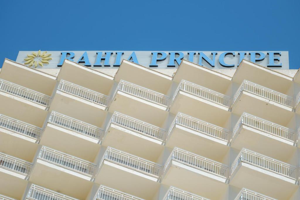 Viesnīcas "Bahia Principe" nosaukums redzams uz ēkas jumta, kas ir balta un mūsdienīga, ar balkoniem. Zila debess veido kontrastu ar viesnīcas fasādi.