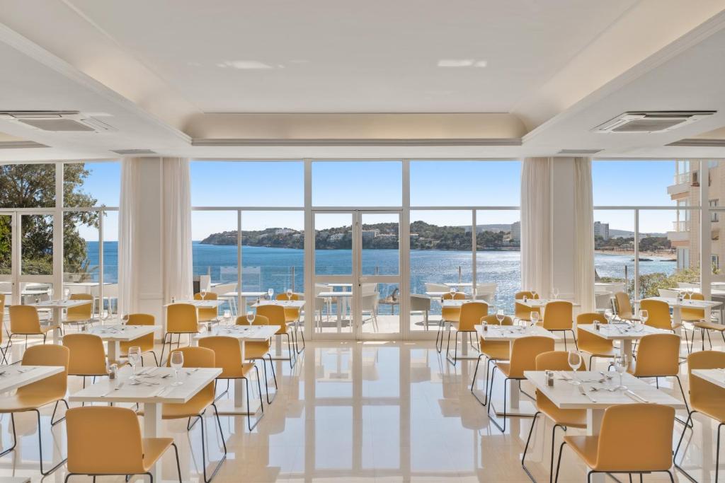 Gaiša un moderna restorāna telpa ar skatu uz jūru, kuras interjerā dominē dzeltenas krēsli. Galdi ir eleganti klāti, radot patīkamu atmosfēru.