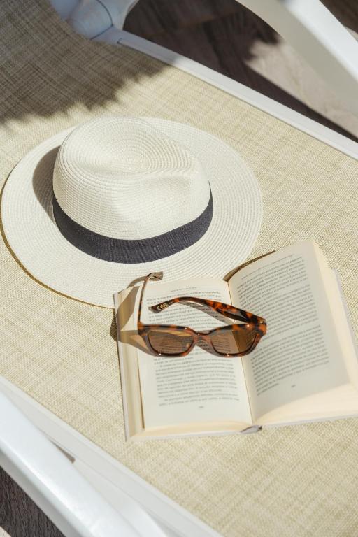 Saulainā pludmales atmosfērā redzams balts cepure ar tumšu joslu, brūnas saulesbrilles un atvērta grāmata uz pludmales krēsla.
