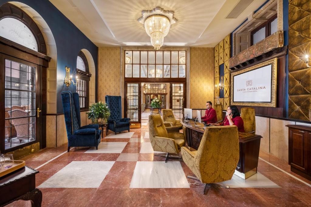 Elegantais viesnīcas reģistratūras iekārtojums ar zeltītiem krēsliem un greznu apgaismojumu. Viesnīcas nosaukums "Santa Catalina" redzams uz lielā ekrāna, radot viesmīlīgu atmosfēru.