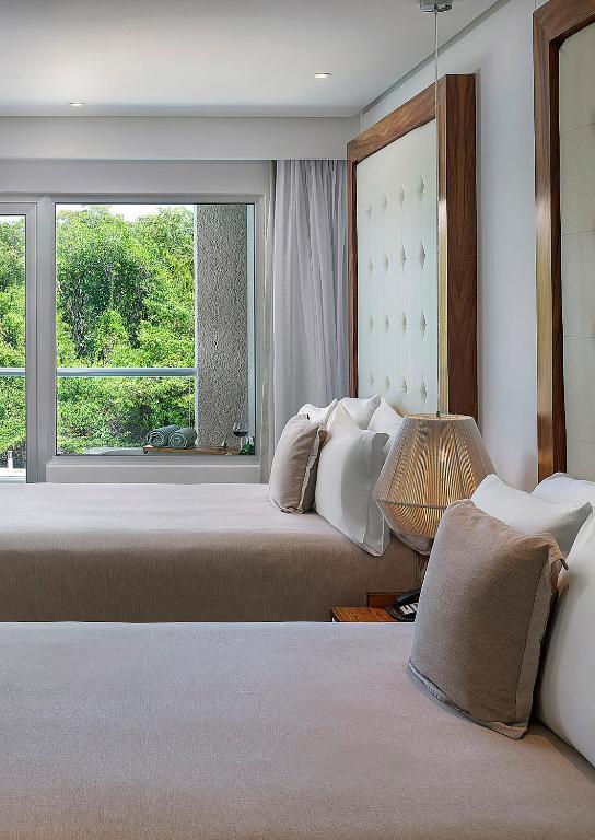 Mūsdienīgs guļamistabas interjers ar divām gultām, lieliem logiem un dabas skatu. Telpa ir gaiša un eleganta, ar stilīgu apgaismojumu un komfortablām spilveniem.