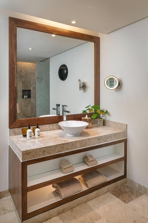 Mūsdienīgs vannas istabas interjers ar elegantu koka izlietni, marmora virsmu un lielu spoguli. Uz letes izvietoti kosmētikas līdzekļi un zaļš augs, radot relaksējošu atmosfēru.