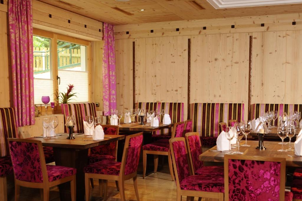 Elegantā restorāna interjers ar koka sienām, rozā aizkariem un krāsainām krēslām. Galdi ir saklāti ar traukiem un glāzēm, radot viesmīlīgu atmosfēru.