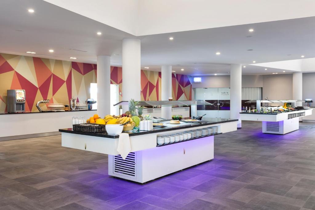 Mūsdienīgs bārs ar dažādu augļu un ēdienu piedāvājumu, kurā redzami gaiši toņi un ģeometriski raksti uz sienas. Telpa ir plaša un labi apgaismota, radot patīkamu atmosfēru.