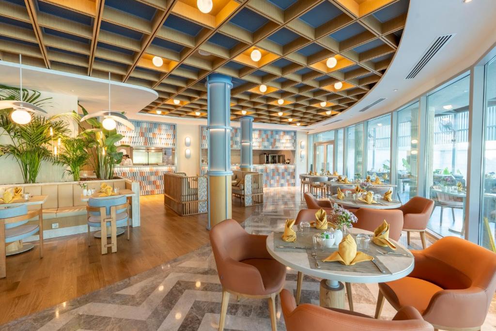 Mūsdienīgs restorāna interjers ar stilīgu dizainu, kurā ir koka griesti un plašas logu sienas, kas ļauj ieplūst dabiskajai gaismai. Galdi ir eleganti klāti ar dzeltenām salvetēm un ziedu kompozīcijām.