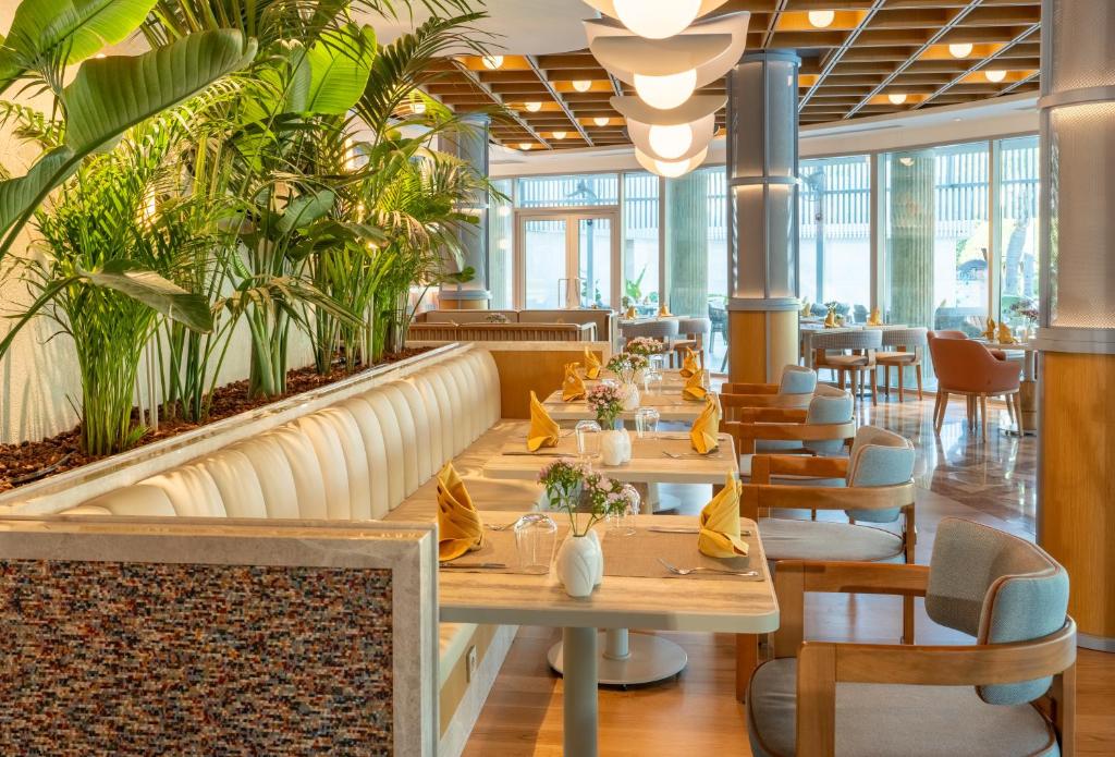 Mūsdienīgs restorāna interjers ar augiem un elegantu galdu uzstādījumu, kas rada patīkamu atmosfēru. Gaisma un dizains piešķir telpai siltumu un komfortu.