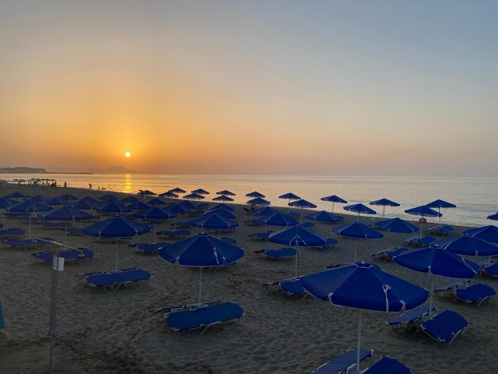 Saulriets pludmalē ar zilām saulessargiem, kas izkārtoti pa smiltīm, radot mierīgu un relaksējošu atmosfēru. Ūdens ir mierīgs, un debesis ir nokrāsotas siltos toņos.