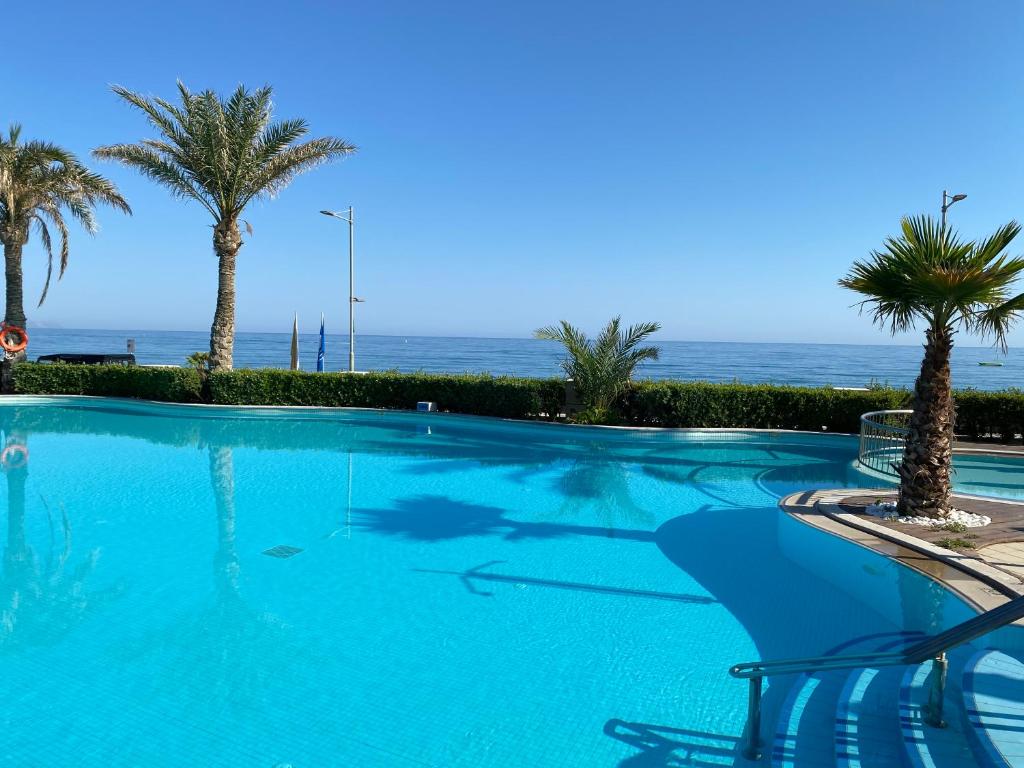 Lieliska skats uz baseinu ar palmām, kas atrodas blakus jūrai. Skats ir skaidrs un saulains, radot relaksējošu atmosfēru.