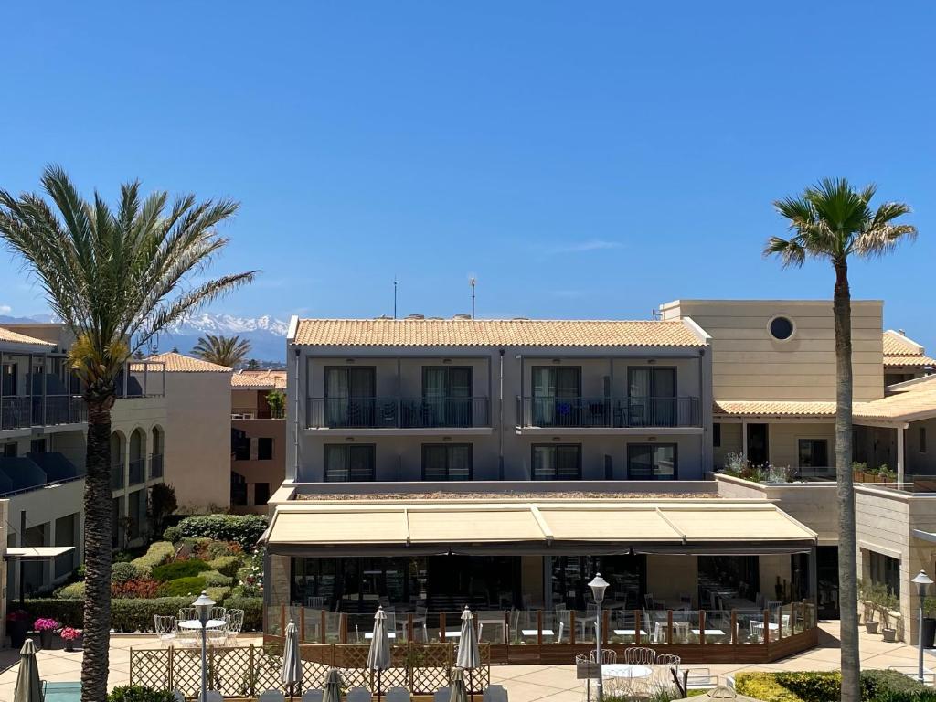 Vasaras kūrorts ar palmām un modernu arhitektūru, kurā redzama viesnīca ar terasi un sniegotiem kalniem fona.