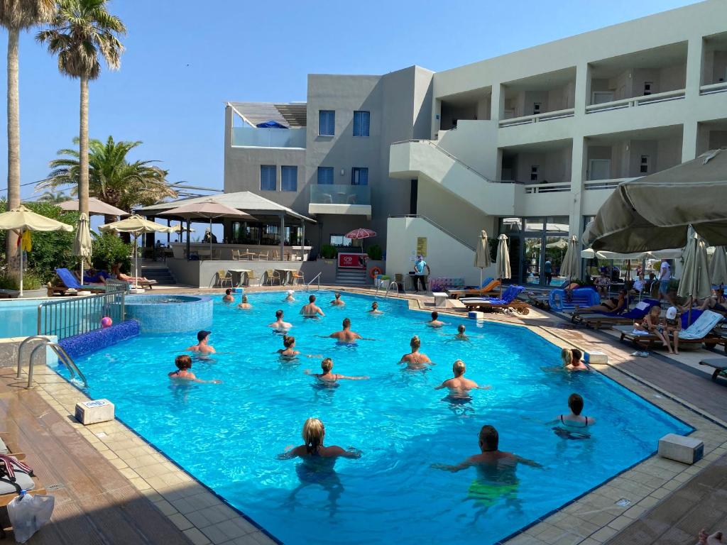 Baseins ar cilvēkiem, kas nodarbojas ar ūdens aerobiku, apkārt palmām un viesnīcas ēkai. Atpūtas zona ir piepildīta ar sauļošanās krēsliem.