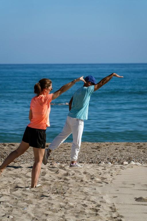 Divi cilvēki veic vingrošanu pludmalē, baudot sauli un jūras tuvumu. Viens no viņiem veic dinamisku kustību, kamēr otrs seko līdzi.