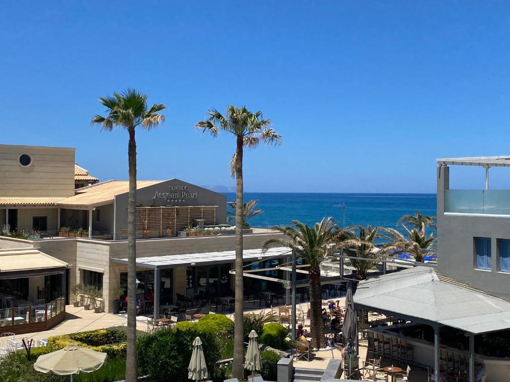 Tropu kūrorta ainava ar palmām un skatu uz jūru, kur redzama ēka ar nosaukumu "Sentido Aegean Pearl". Vasaras atmosfēra un relaksējoša vide.