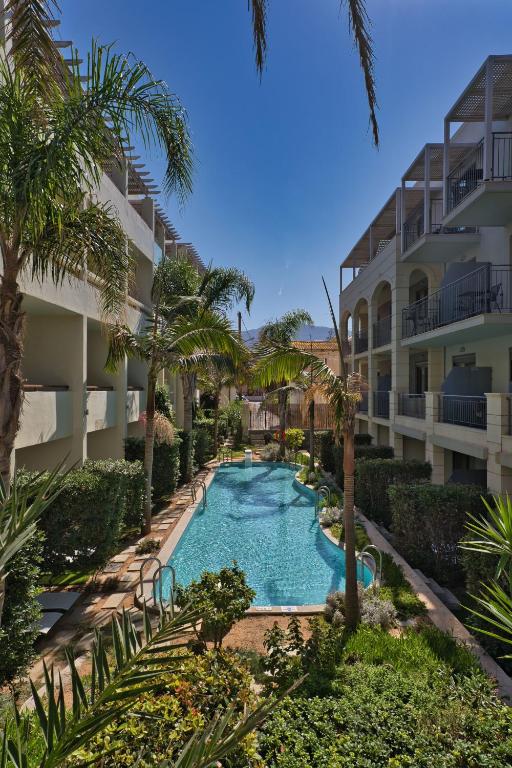 Skaista viesnīcas teritorija ar peldbaseinu, kas ieskauta zaļām palmām un augiem. Dārzā redzamas labi koptas ainavas un ēkas ar balkoniem.