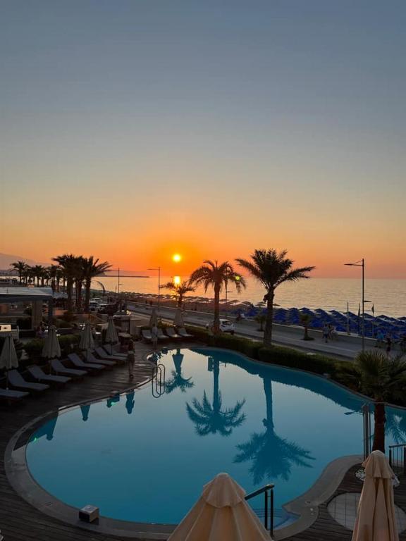 Saulriets pie baseina, kur atspoguļojas palmu silueti un jūra, radot mierīgu un gleznainu ainavu.