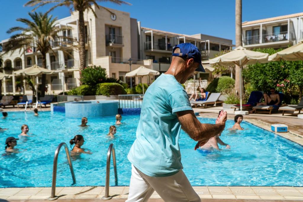 Vīrietis peldbaseina malā, baudot sauli un skatoties uz apmeklētājiem, kuri peld baseinā. Apkārt ir palmām ieskauts viesnīcas komplekss ar atpūtas krēslām.