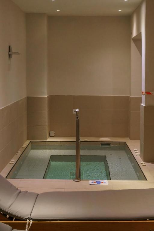 Mūsdienīgs spa telpa ar mazāku baseinu, kurā ir ūdens burbuļi un metāla stabs centrā. Telpa ir gaiša un eleganta, radot relaksējošu atmosfēru.