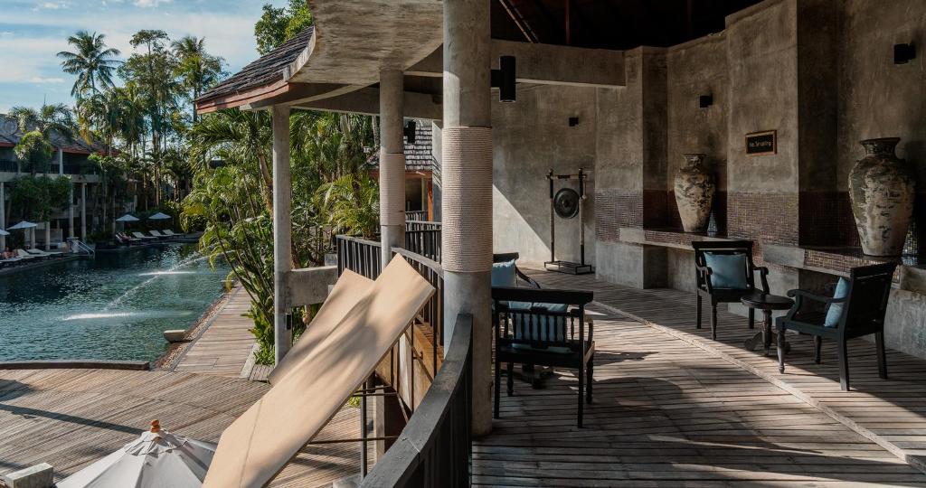 Relaksējoša atpūtas zona ar skatu uz baseinu, kurā ir koka terase, krēsli un rotājumi. Apkārt augstas palmas un pludmales gultas.