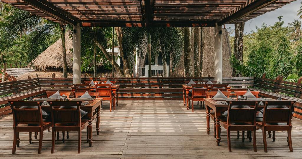 Ārtelpa restorānā ar koka galdiem un krēsliem, ierīkota zem jumta, ar skatu uz zaļiem kokiem un dabu. Ideāla vieta relaksācijai un maltītēm brīvā dabā.