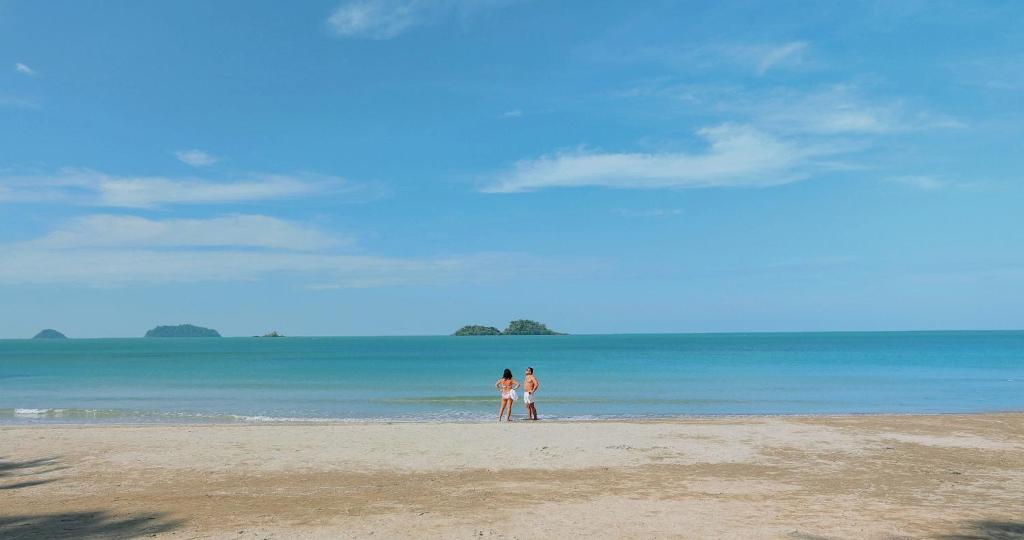 Divas personas stāv uz pludmales, skatoties uz mierīgu jūru un tālu salām, fons ar skaidru, zilu debesu un viegliem mākoņiem.